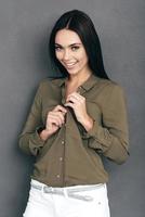 Unbuttoning her shirt. Attractive young woman unbuttoning her shirt and looking at camera with smile while standing against grey wall photo