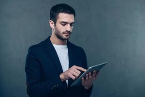 Have some work to do. Handsome young man working on his digital tablet while standing against grey background photo