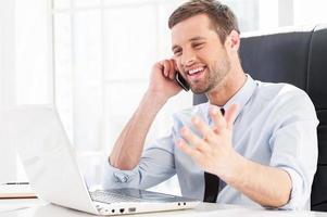 Happy with business going. Happy young man in formalwear looking at computer monitor and talking on the mobile phone while sitting at his working place photo