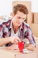 Packing boxes. Handsome young man packing boxes while other cardboard boxes laying in the background photo