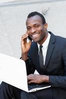 Living a business life. Cheerful young African man in formalwear talking on the mobile phone and working on laptop while sitting outdoors photo