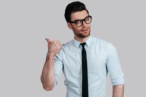 Pointing copy space.  Handsome young man looking at camera and pointing away while standing against grey background photo