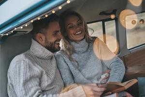 gran historia. hermosa pareja joven leyendo el libro y sonriendo mientras se sienta en una mini furgoneta de estilo retro