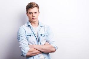 Young and confident. Handsome young man keeping arms crossed while standing against grey background. photo