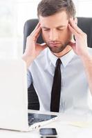 sentirse cansado y deprimido. joven deprimido con camisa y corbata sosteniendo la cabeza en las manos y manteniendo los ojos cerrados mientras se sienta en su lugar de trabajo foto