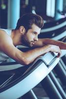 Feeling tired. Side view of young man in sportswear looking exhausted while leaning on treadmill at gym photo