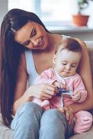 La mejor mamá. bella joven sosteniendo a una niña con un juguete de rodillas y mirándola con una sonrisa mientras se sienta en el sofá de casa foto