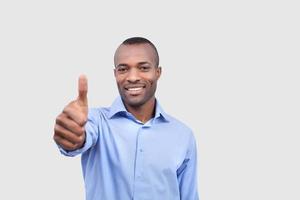 Everything is all right. Cheerful young black man smiling at camera and stretching out his hand with thumb up while isolated on grey background photo