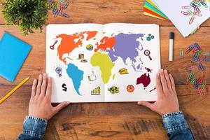 Planning his journey. Top view close-up image of man holding hands on his notebook with colorful map on it while sitting at the wooden desk photo