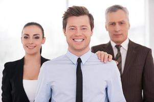 Real team leader. Cheerful young man in formalwear looking at camera and smiling while his colleagues standing behind her photo