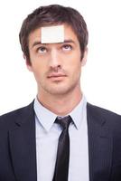 Things to do. Portrait of frustrated young man in formalwear and adhesive note on his forehead standing against white background photo