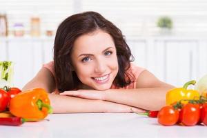 chica vegetariana. atractiva mujer joven apoyada en la mesa de la cocina y sonriendo mientras verduras coloridas yacen cerca de ella foto