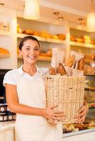 el pan más fresco para nuestros clientes. Hermosa mujer joven en delantal sosteniendo una cesta con pan y sonriendo mientras está de pie en la panadería. foto