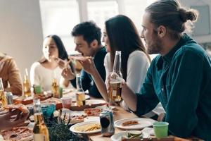 Great time with friends. Group of young people in casual wear eating pizza and smiling while having a dinner party indoors photo