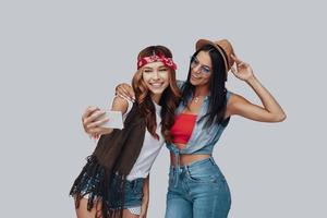 Two attractive stylish young women taking selfie and smiling while standing against grey background photo