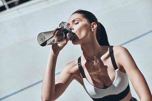 mantenerse hidratado. bella joven con ropa deportiva bebiendo agua mientras se sienta en la pista de atletismo al aire libre foto