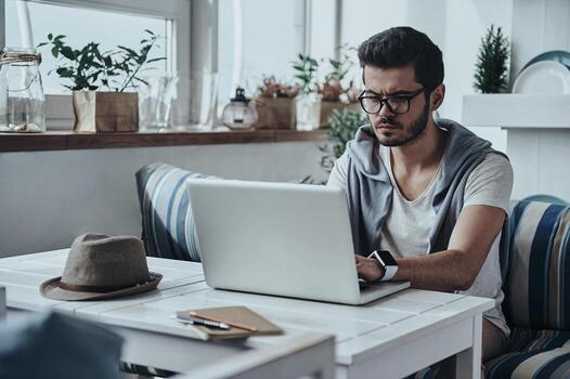 Putting ideas into something real. Handsome young modern man in eyewear using computer while sitting on the sofa photo
