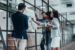 Searching for right decision. Group of confident people planning business strategy while young woman pointing at large paper displayed on the glass wall in the office hallway photo