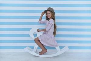 Cute little girl adjusting her hat and looking at camera with smile while sitting on the toy horse photo