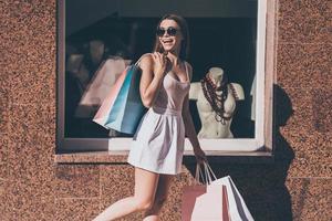 hermosa adicta a las compras. hermosa joven que lleva bolsas de compras y mira por encima del hombro con una sonrisa mientras camina por la calle con una tienda de moda en el fondo foto