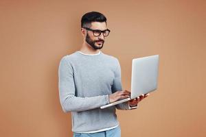 Busy young man in smart casual wear using laptop while standing against brown background photo