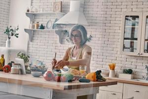 Busy senior woman in apron cooking healthy dinner while spending time at home photo