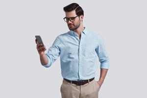 More than useful device.  Good looking young man using smart phone and keeping hand in pocket while standing against grey background photo