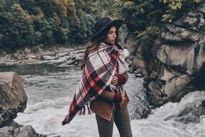 mujer con estilo. atractiva mujer joven cubierta con una manta mirando hacia otro lado mientras está de pie cerca del río en las montañas foto