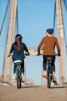 Enjoying their ride together. Rear view of beautiful young couple riding bicycles along the bridge and looking at each other photo