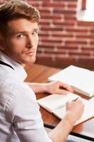 Writing his new roman. Top view of handsome young man in shirt and tie writing something in note pad and looking at camera while sitting at his working place photo