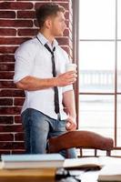 Refreshing his mind. Handsome young man in shirt and tie holding coffee cup and looking through the window while sitting at the window sill photo