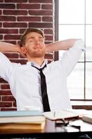 tomando tiempo para relajarse. un joven apuesto con camisa y corbata sosteniendo los ojos cerrados y tomándose de la mano detrás de la cabeza mientras estaba sentado en su lugar de trabajo foto