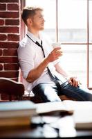 Refreshing his mind with cup of coffee. Handsome young man in shirt and tie holding coffee cup and looking through the window while sitting at the window sill photo