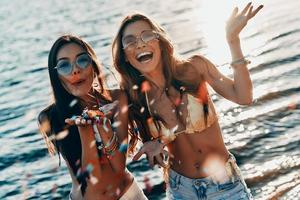 divertido con la mejor amiga. vista superior de dos atractivas mujeres jóvenes en traje de baño sonriendo y soplando confeti mientras están de pie cerca del río al aire libre foto