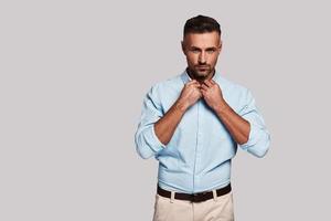 Great sense of style. Handsome young man adjusting collar and looking at camera while standing against grey background photo