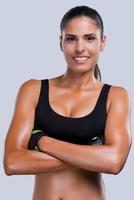 Fit and beautiful. Beautiful young sporty woman in sports clothing keeping arms crossed and smiling while standing against grey background photo