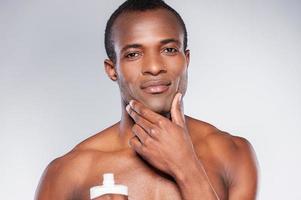 No skin irritation. Young shirtless African man applying cream at his face and looking at camera while standing against grey background photo