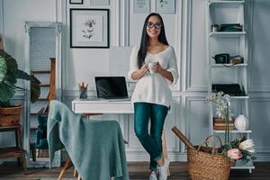 Coffee brings fresh ideas. Beautiful young woman looking at camera and smiling while standing in home office photo