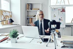 hombre mayor con camisa elegante y corbata trabajando usando una computadora portátil mientras hace videos en las redes sociales foto