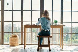 Rear view of young woman in smart casual wear using laptop while sitting in front of the window photo