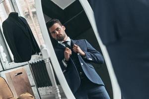 Confident in his style. Reflection of handsome young man in full suit adjusting his jacket while standing in front of the mirror indoors photo