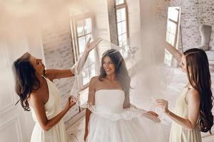 Enjoying wedding preparations. Top view of bridesmaids helping bride to put on a veil and smiling while standing in the fitting room photo