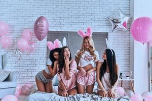 Effortless beauties. Four playful young smiling women in bunny ears making a face and gesturing while sitting on the bed with balloons around photo