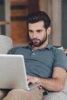 Modern blogger. Handsome young man using his laptop while sitting on the couch at home photo