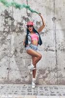 Hot and funky beauty. Full length of playful young women holding smoke bomb and smiling while posing against the concrete wall photo