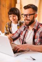 Sharing an important experience. Smiling young man working on laptop with his son while sitting at the working place photo