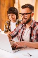 compartiendo una experiencia importante. un joven sonriente que trabaja en una laptop con su hijo mientras está sentado en el lugar de trabajo foto