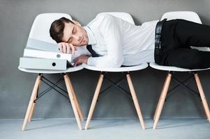 Tired after hard working. Tired young man keeping eyes closed while laying on chairs against grey background photo
