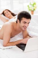 Surfing web in bed. Cheerful young man using laptop while lying in bed with his girlfriend photo