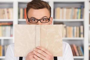 Shocking story. Surprised young nerd man in glasses looking out of the book while standing at the library photo